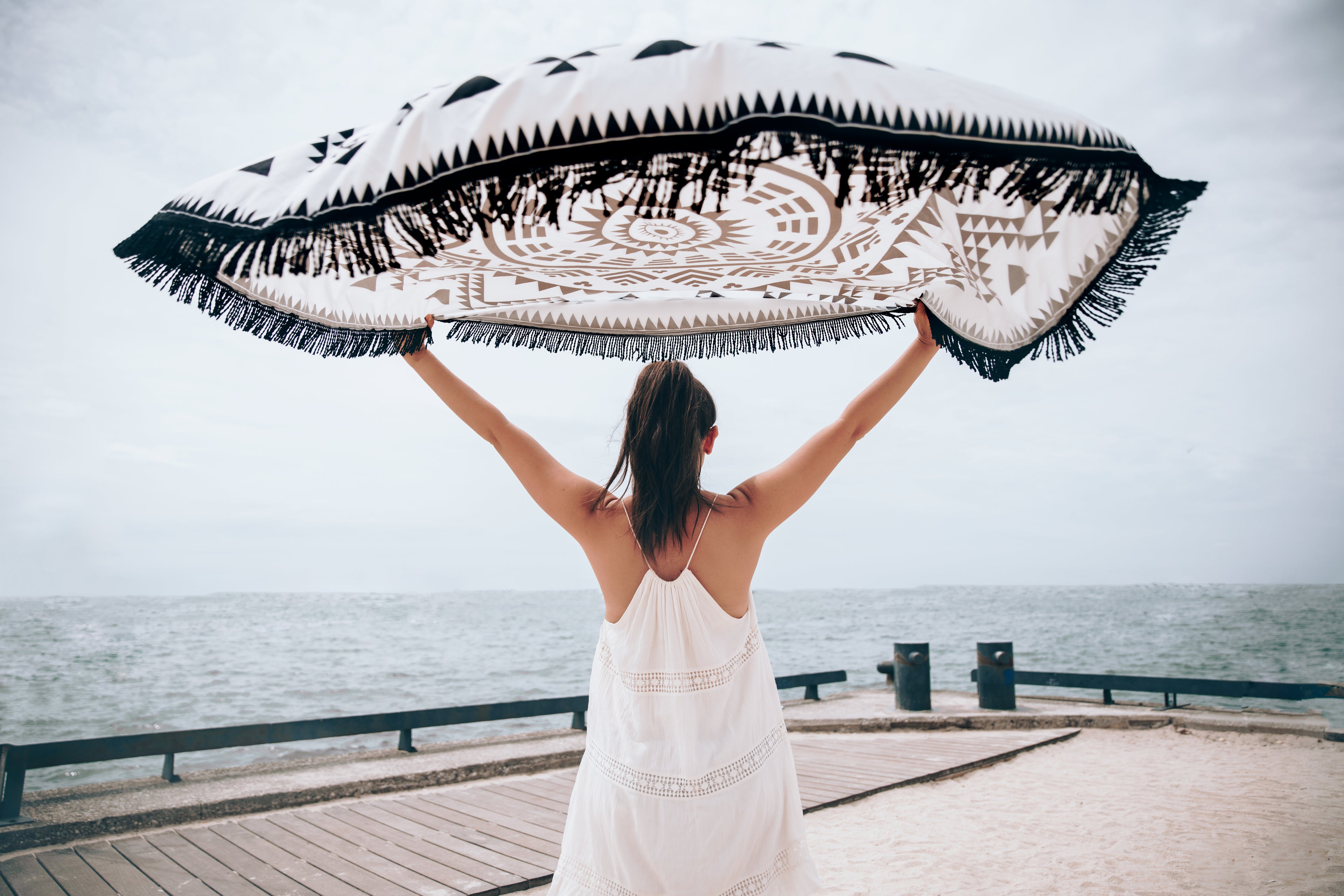 Une jeune fille en robe blanche tient au-dessus de sa tête une serviette de plage avec un motif original. 