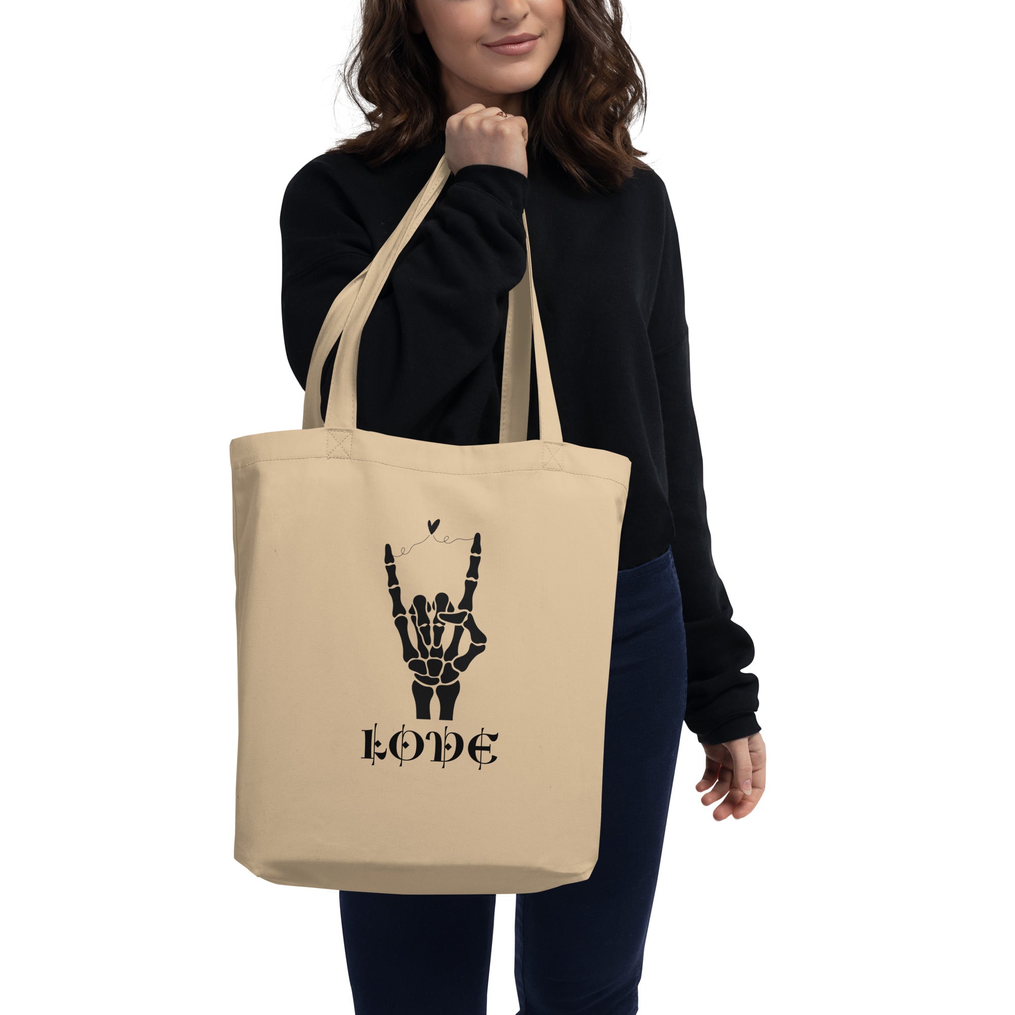 Un tote bag en toile de couleur beige naturel présentant une illustration noire centrée. Le dessin représente une main de squelette faisant le signe des cornes (rock on) avec un petit cœur flottant au-dessus des doigts. En dessous, le mot "LOVE" est inscrit dans une police de caractères stylisée, médiévale et gothique.